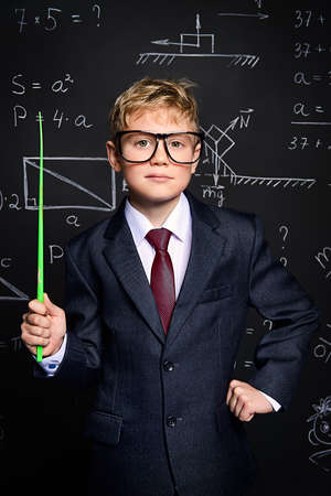 Smart schoolboy in black suit and glasses standing by a school blackboard. Educational concept.の写真素材