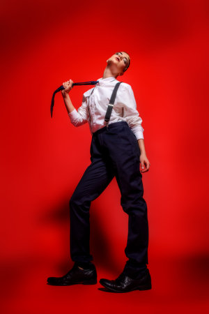 Fashion shot. Attractive young woman posing in a man's shirt and trousers with suspenders. Man's style clothing. Red background.の写真素材