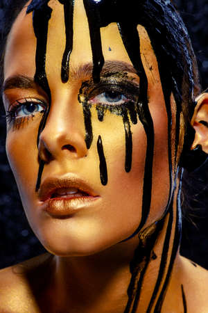 Beauty provocation. Body painting project. Portrait of a young woman with golden skin and black oil pouring on her. Fashionable provocation. Pollution and poisoning concept.の写真素材