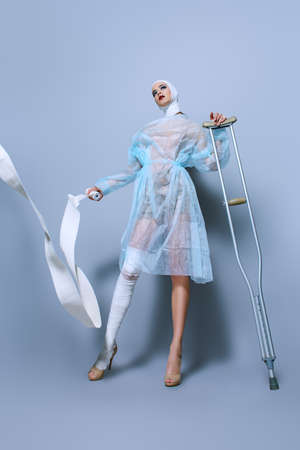 Fashion shot. Gorgeous female model in bandages and hospital gown posing at studio with crutches. Beauty and medicine, plastic surgery.の写真素材