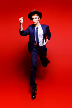 Full length portrait of an attractive young woman posing in a man's suit and a hat. Man's style clothing. Red background. Fashion shot.の写真素材