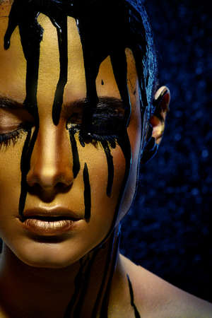 Beauty provocation. Body painting project. Portrait of a young woman with golden skin and black oil pouring on her. Fashionable provocation. Pollution and poisoning concept.の写真素材