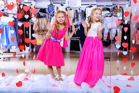 Two cute little girls in beautiful dresses posing in the window of children's clothing store. Kid's fashion. Seasonal sale and shopping.の写真素材