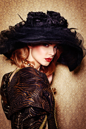 Portrait of a gorgeous young woman in elegant broad-brimmed hat and luxurious dress over golden...