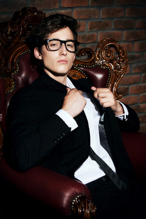 Glamorous sexy young man in elegant black suit and glasses sitting in vintage armchair. Male beauty,...