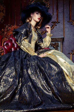 Portrait of a beautiful young woman in the lush expensive dress and elegant broad-brimmed hat in an...