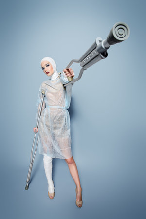 Fashion shot. Gorgeous female model in bandages and hospital gown posing at studio with crutches....