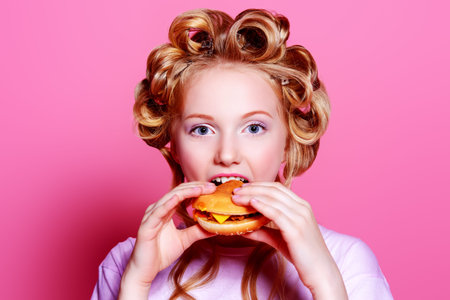 Pretty teen girl with curlers on her hair is eating a hamburger. Pink background. Beauty, fashion...