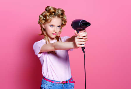 Portrait of a pretty girl teenager with curlers in her blonde hair holding hair dryer. Teen style, fashionable teen girl. Studio shot over pink background.の写真素材