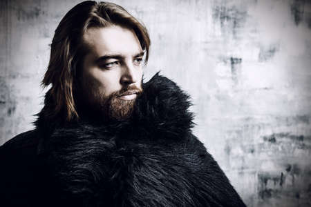Fashion shot of a stylish brutal bearded man wearing fur coat. Studio shot over grunge background.の写真素材