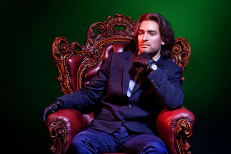 Handsome man in an expensive suit and black leather gloves sitting in a classical vintage armchair. Male beauty, fashion. Accessories.の写真素材