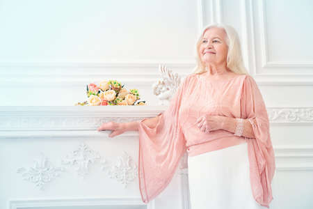 Modern well-groomed old woman with beautiful blonde hair standing in a room with a classic interior. Beauty, fashion. Luxury.の写真素材