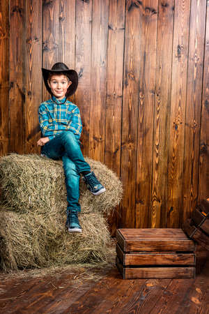 Portrait of a cute nine year old boy posing near the hay on the background of wooden wall. Western style, cowboy. Kid's fashion. Clothes for children.の写真素材