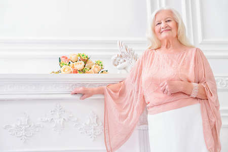 Modern well-groomed old woman with beautiful blonde hair standing in a room with a classic interior. Beauty, fashion. Luxury.の写真素材