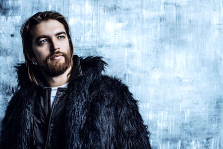 Fashion shot of a stylish brutal bearded man wearing fur coat. Studio shot over grunge background.の写真素材