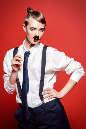 Fashion shot. Attractive young woman posing in a man's suit and mustache. Man's style clothing. Red background.の写真素材