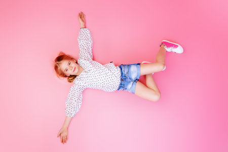 Happy childhood. Cute eight year old girl wearing summer jeans shorts and blouse lying on a pink...