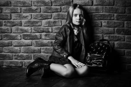 Black-and-white portrait of a calm cute girl sitting by the brick wall. Urban style clothes. Children's fashion.の写真素材