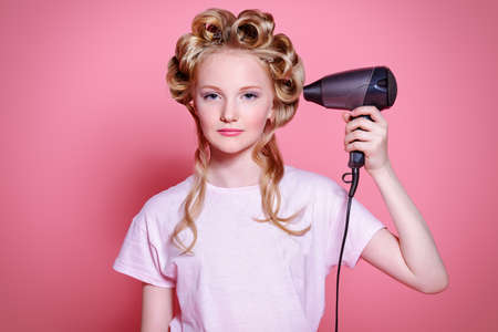 Portrait of a pretty girl teenager with curlers in her blonde hair holding hair dryer. Teen style, fashionable teen girl. Studio shot over pink background.の写真素材