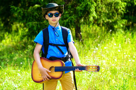 Modern ten year old boy goes hiking with a guitar and a backpack. Sunny summer holidays.の写真素材