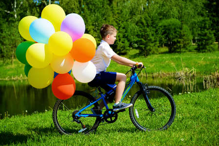 Happy boy is riding a bicycle with colored balloons in a park. Summer holidays. Birthday.の写真素材