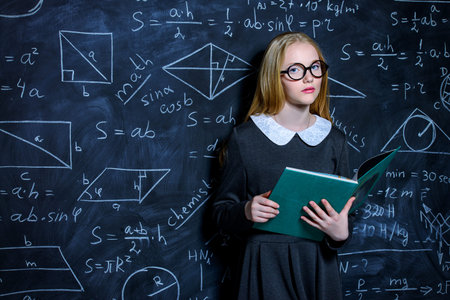 Education. Smart student girl wearing black school dress standing by a blackboard with a book....