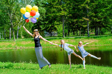 Happy summer day. Cheerful mother runs with her little daughters with balloons through the park. Family concept.の写真素材