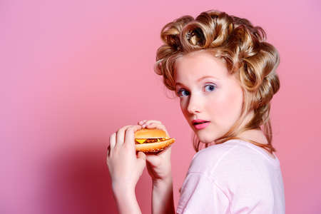 Pretty teen girl with curlers on her hair is eating a hamburger. Pink background. Beauty, fashion concept.の写真素材