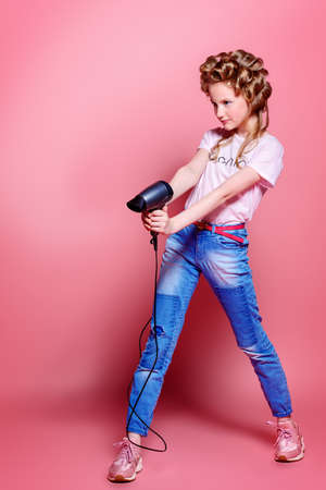 Portrait of a pretty girl teenager with curlers in her blonde hair holding hair dryer. Teen style, fashionable teen girl. Studio shot over pink background.の写真素材