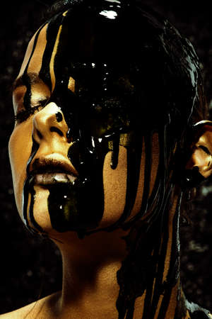 Beauty provocation. Body painting project. Portrait of a young woman with golden skin and black oil pouring on her. Fashionable provocation. Pollution and poisoning concept.の写真素材