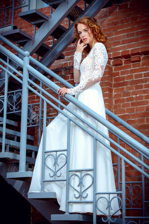 Beautiful young woman with bright foxy hair wearing evening white dress on a city street. Charming bride woman posing outdoor. Urban fashion.の写真素材