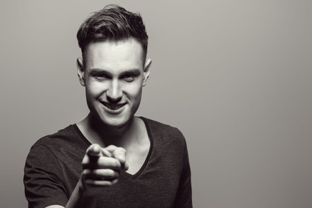 Black-and-white portrait of a smiling man pointing his index finger at the camera. Studio shot. Copy...