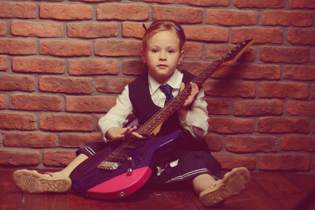 Modern little girl in school uniform posing with electric guitar over brick wall background. Rock star, rock music, pop music concept.の写真素材