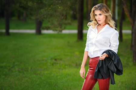 Fashionable gorgeous woman wearing red leather trousers and white blouse posing at a park.