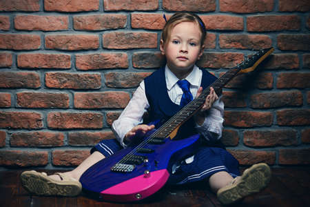 Modern little girl in school uniform posing with electric guitar over brick wall background. Rock star, rock music, pop music concept.の写真素材