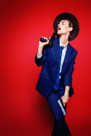 Portrait of an attractive young woman posing in a man's suit and a hat with guns. Gangster style. Man's style clothing. Red background. Fashion shot.の写真素材