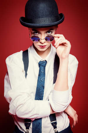 Fashion shot. Attractive young woman posing in a man's clothes and bowler hat. Man's style clothing. Red background.の写真素材
