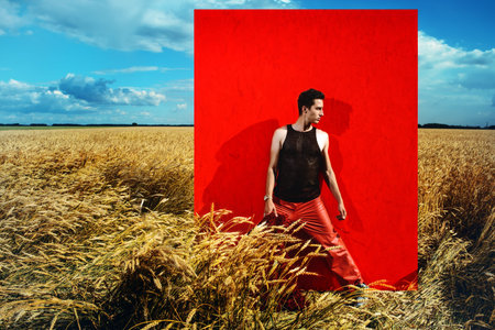 Fashion shot of a professional handsome male model in long red fur coat posing in a wheat field....