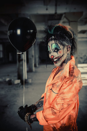 Sinister clown man stained in blood is standing in an abandoned house with a balloon. Male zombie...