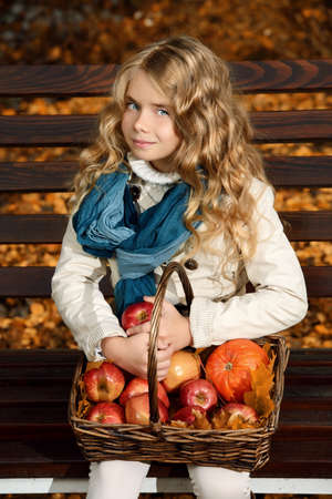 Lovely little girl sitting on a bench with a basket of red apples in a beautiful autumn park. Children's fashion.の写真素材