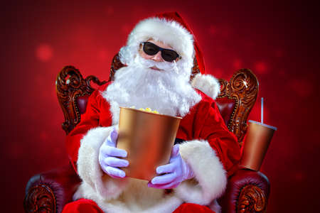 Merry Christmas and Happy New Year. Santa Claus sitting on his armchair and eating popcorn and drinking soda. Entertainment and cinema concept. Red background.の写真素材