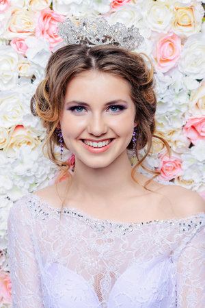 Charming bride in a beautiful dress and a crown posing by a background of roses. Wedding fashion.