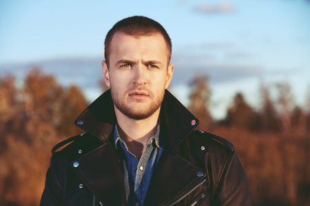 Handsome young man in leather jacket in the autumn park on a sunset. Seasonal fashion.
