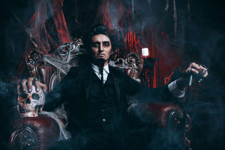 Halloween. Frightening gloomy man in a black tailcoat sits in an armchair with a skull in an old abandoned castle. The Dark Lord. Vampire man.の写真素材