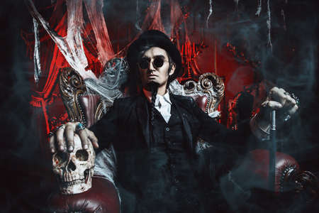 Halloween. Frightening gloomy man in a black tailcoat sits in an armchair with a skull in an old abandoned castle. The Dark Lord. Vampire man.の写真素材