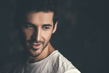 Portrait of a handsome smiling young man wearing white t-shirt. Studio shot over dark background. Men's beauty and health.の写真素材