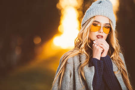 Autumn fashion. Fashionable young woman wearing warm sweater, a coat and a woolen hat posing in the park at a sunset.の写真素材