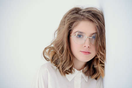 Portrait of a cute girl teenager in spectacles leaned against the white wall. Glasses for a modern young generation. Beauty, fashion.の写真素材