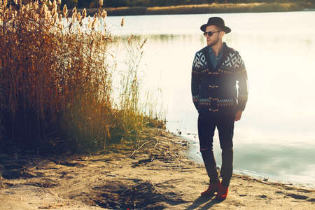 Handsome man wearing cardigan and hat stands outdoor over autumn nature background. Autumn fashion.の写真素材