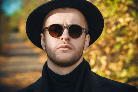 Seasonal autumn fashion. Close-up portrait of a handsome man wearing black hat and sunglasses in the autumn park.の写真素材